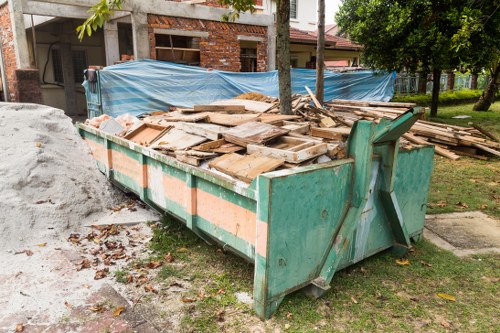 A large industrial skip container filled with broken wooden pallets, in an outdoor area on a residential street. The pallets are weathered, with some showing chipped edges and splintered surfaces, and are stacked haphazardly within the green-painted metal skip, which has orange reflective stripes and visible wear on its surface. The skip is positioned on a gravel and dirt surface, with scattered leaves and small debris around its base. In the background, there is a partially constructed brick building covered with blue tarpaulin and construction materials, along with a tree providing some greenery. The surrounding environment suggests ongoing building or renovation work, consistent with rubbish removal services by Garden Clearance Waterloo, serving the Waterloo area in London. Natural daylight illuminates the scene, highlighting the textures of the wood and the weathered finish of the skip.