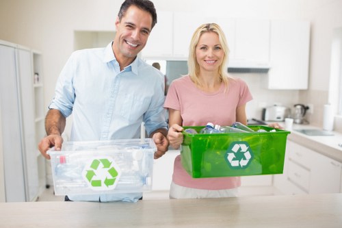 A man and a woman standing in a modern kitchen holding green and clear recycling bins with the recycling symbol visible on each. The man, wearing a light blue shirt, holds a clear bin filled with plastic bottles and packaging, while the woman, dressed in a pink top, holds a larger green bin. The kitchen features white cabinetry, a white countertop, and kitchen appliances in the background, with natural light illuminating the scene. The image reflects an environmentally conscious approach to rubbish and waste management, aligning with services provided by Garden Clearance Waterloo, operating within Waterloo, London, and nearby postcode areas.