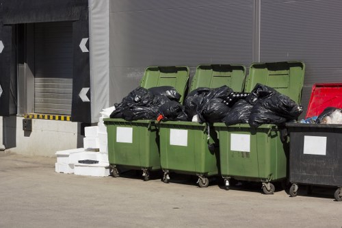 Four large wheeled rubbish bins are positioned in a row on a paved outdoor area, with the first three bins being green and the fourth bin black. The green bins have their lids open, revealing black plastic refuse bags piled inside, some with visible red ties. The black bin appears to contain additional waste, possibly plastic or general rubbish. Behind the bins, there is a building with grey metallic cladding and a dark roller shutter door with white directional arrows. To the left of the bins, a small stack of white bricks or concrete blocks is arranged on the ground. The environment appears to be a commercial or industrial location, typical of waste collection or disposal areas, with clear sunlight illuminating the scene, contributing to a bright and neutral atmosphere. This image relates to rubbish collection or clearance services, potentially in the Waterloo area, as indicated by the website domain.