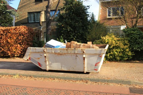 A large white metal skip with visible rust and dirt marks, positioned on a paved driveway in a residential area, containing several cardboard boxes and miscellaneous debris. The skip is located near the curb, adjacent to a brick-paved street, with a background featuring a row of houses with brick and stucco exteriors, windows, and sloped roofs. Surrounding the skip are neatly trimmed bushes and small trees, some with autumn-colored foliage, indicating a suburban environment. The image appears to have been taken during daylight hours, with natural sunlight illuminating the scene, emphasizing the textures of the cardboard, metal, and surrounding greenery. This scene is representative of rubbish removal services provided by Garden Clearance Waterloo in the Waterloo area, focusing on the collection and disposal of household waste and debris from garden or property clearance jobs.